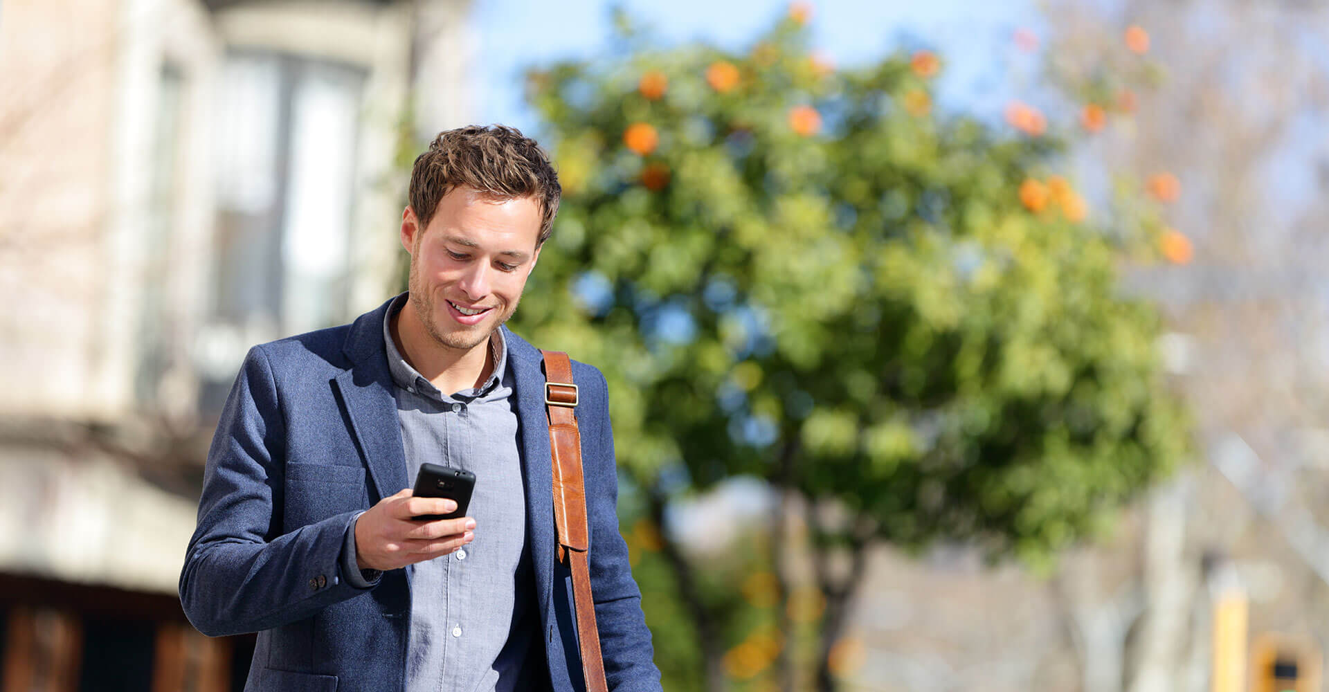 Smiling man in a blazer checking his phone outdoors on a sunny day.