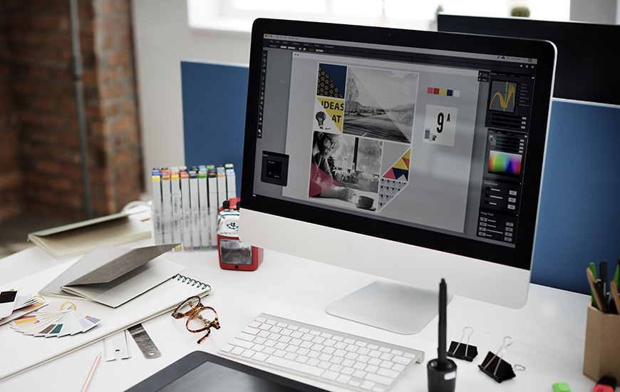 A modern workspace with a computer displaying graphic design software and various art supplies.