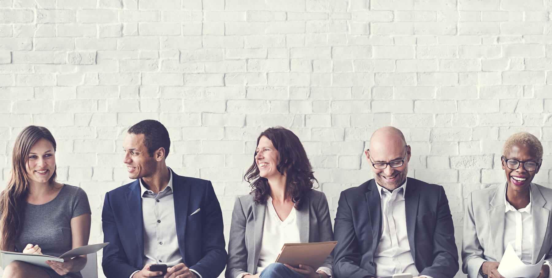 Five professionals sitting against a white brick wall, smiling and holding documents.