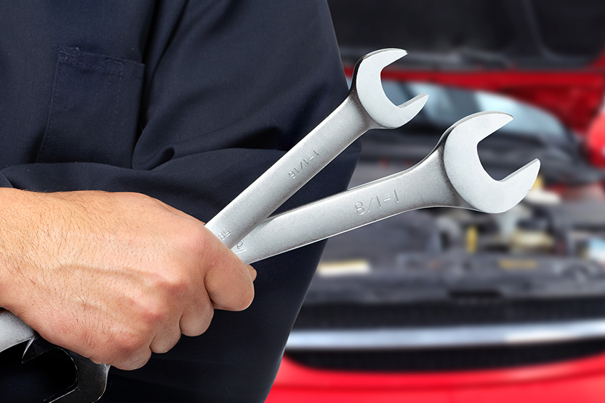 Mechanic holding two wrenches with a car engine in the background.