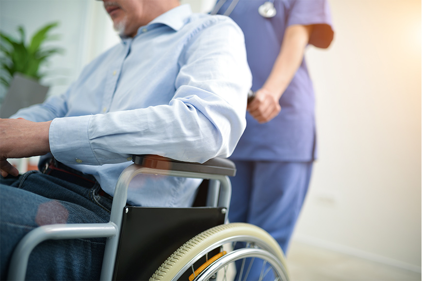 A nurse pushes an elderly man in a wheelchair indoors.