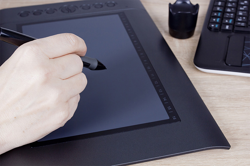 Hand using a stylus on a digital drawing tablet beside a keyboard.