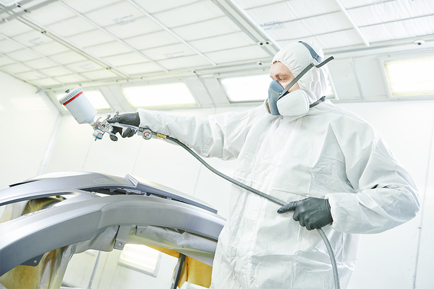 Worker in protective gear spray painting a car part in a bright workshop.