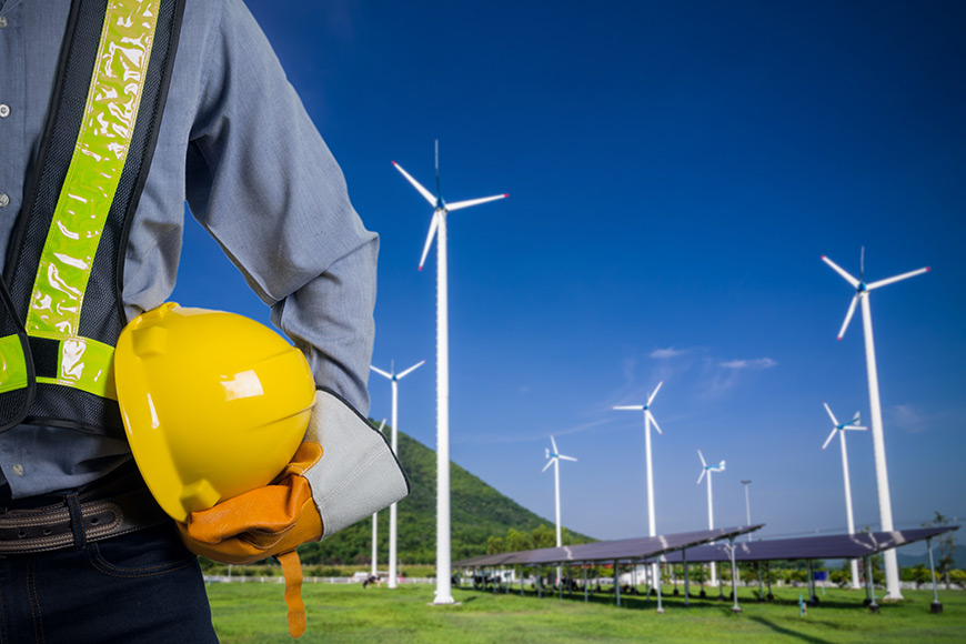 Engineer holding a yellow helmet near wind turbines and solar panels under a clear blue sky.