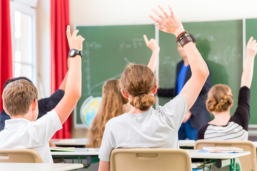 Students raising their hands in a classroom while the teacher stands near the blackboard.