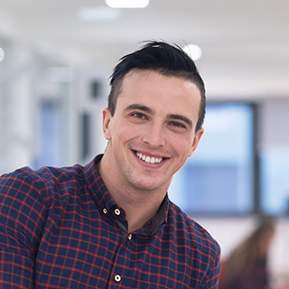 Young man smiling indoors, wearing a checkered shirt.
