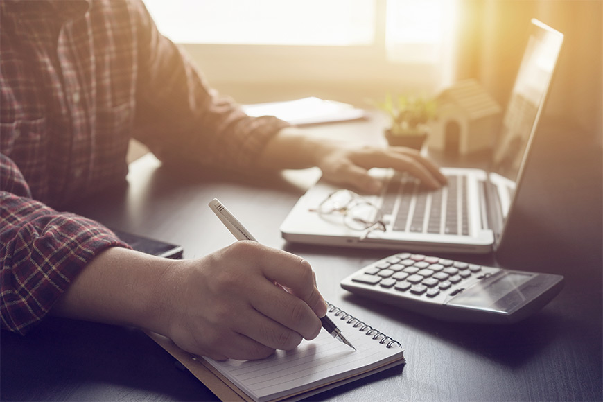 Person writing notes while using a laptop and calculator on a desk.
