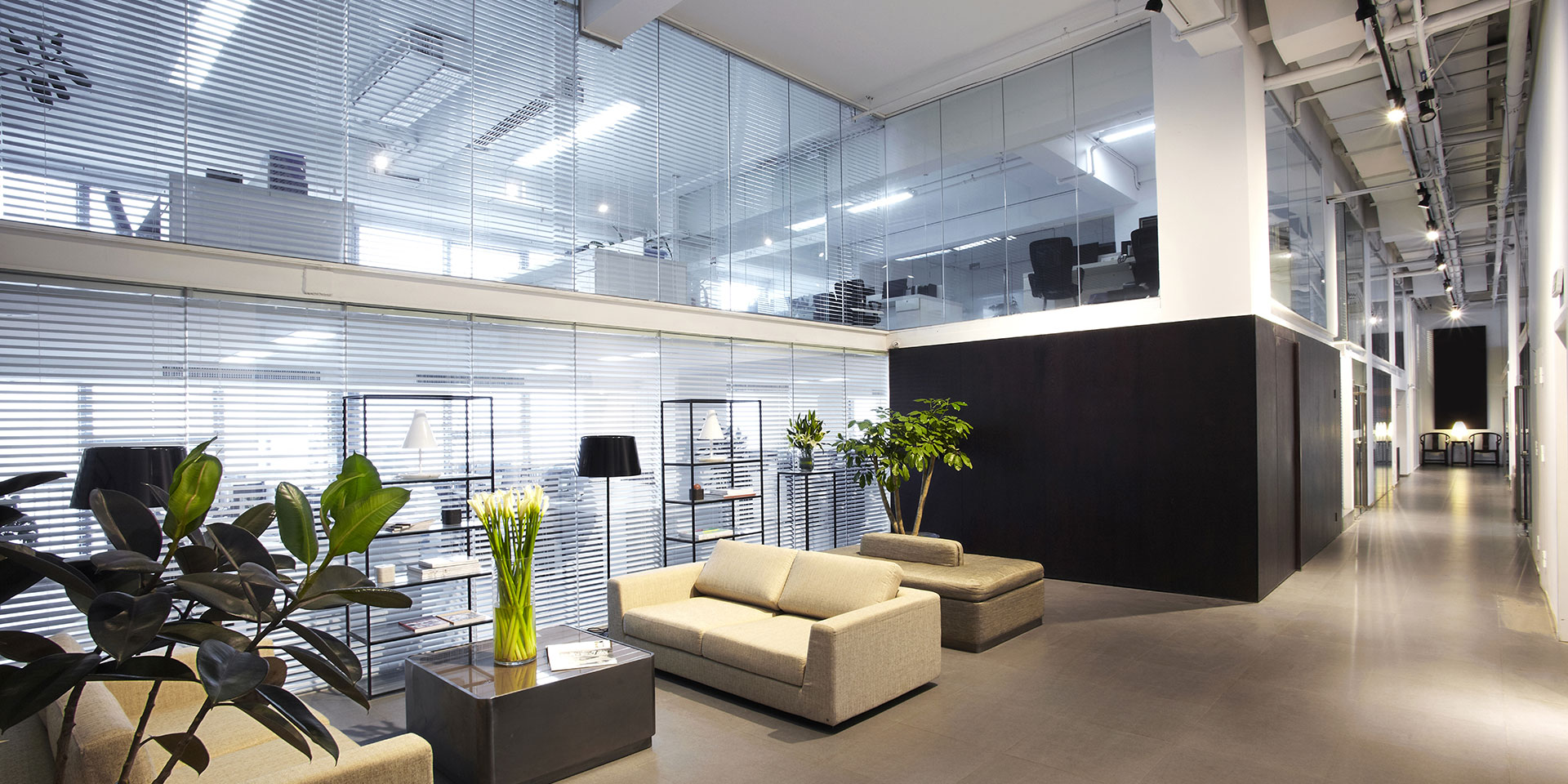 Modern office lounge with beige sofas, plants, and glass walls.