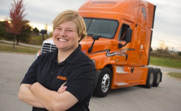 Smiling truck driver stands confidently in front of an orange semi-truck.
